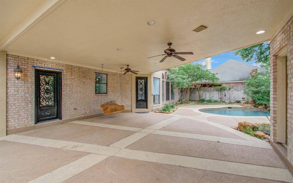 This photo showcases a spacious covered patio with ceiling fans and decorative lighting. It features a swinging seat and opens to a beautifully landscaped backyard with a small pool, surrounded by trees and a privacy fence, perfect for outdoor relaxation and entertainment.