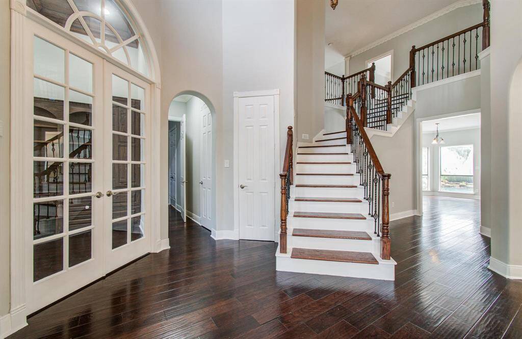 The photo showcases a spacious, elegant foyer with dark hardwood floors, a grand staircase with wooden handrails and iron spindles, and large French doors. The area is bright and open, leading to additional rooms with ample natural light.