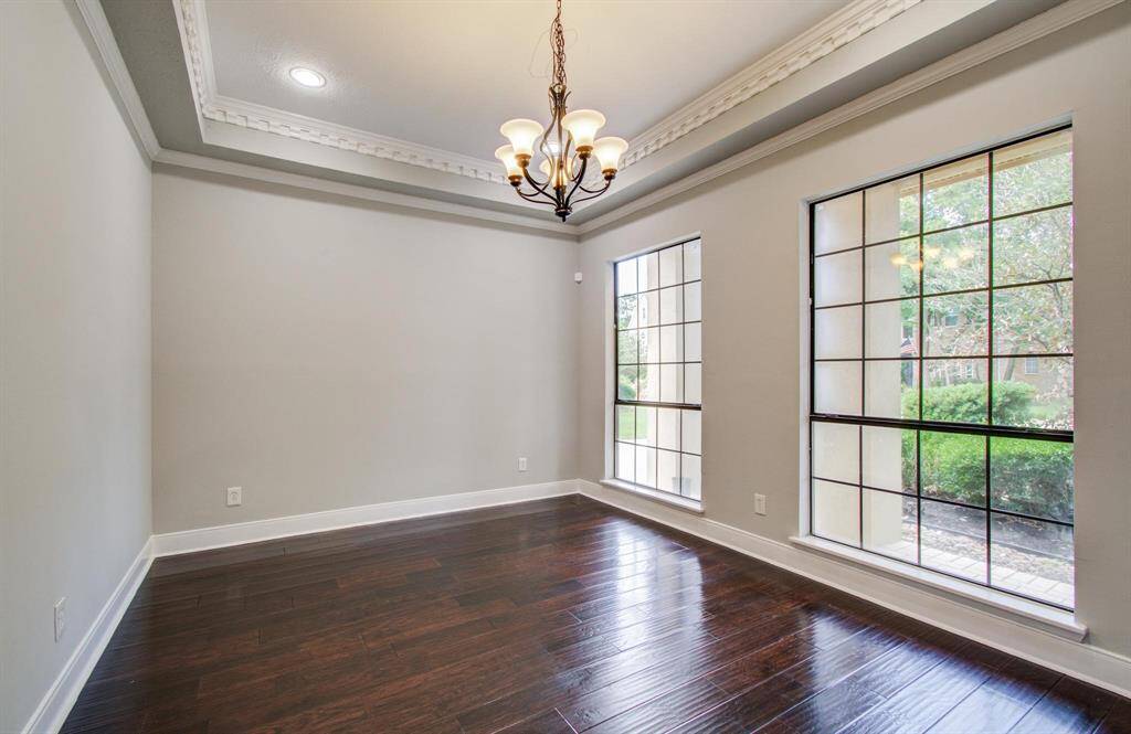 This room features elegant dark hardwood floors, a stylish tray ceiling with crown molding, and large windows offering ample natural light. A classic chandelier provides a warm ambiance, making it perfect for a dining or living space.
