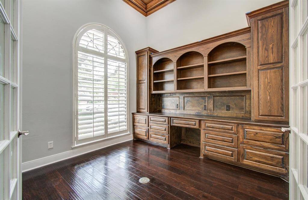 This room features elegant dark wood flooring, built-in wooden shelving and cabinetry, and a large arched window with shutters, providing ample natural light—ideal for a home office or library.