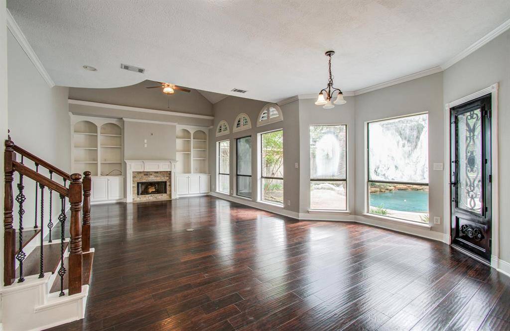 This spacious living area features elegant dark hardwood floors, a cozy fireplace with built-in shelving, and large windows offering picturesque views of a waterfall. The open layout includes a stylish staircase and a decorative front door, creating a welcoming atmosphere.