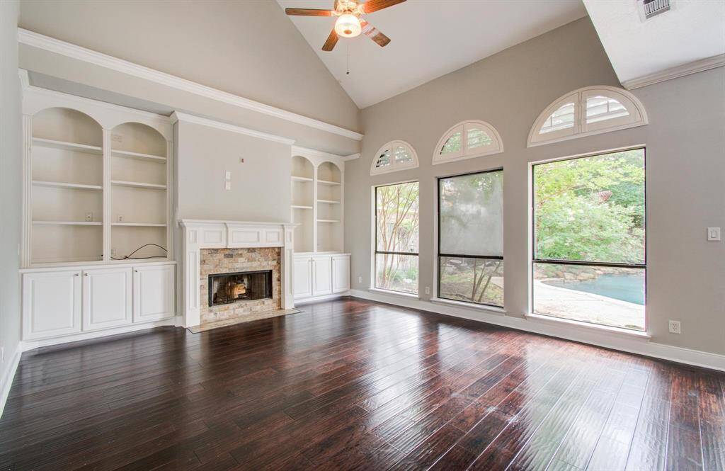 This spacious living room features high ceilings, dark hardwood floors, a cozy fireplace, built-in shelving, and large windows offering abundant natural light and views of the backyard pool.