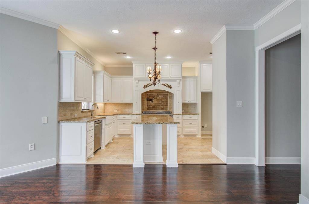 This kitchen features elegant white cabinetry, granite countertops, and a central island. It is accented with a stylish chandelier and has hardwood floors transitioning to tile.