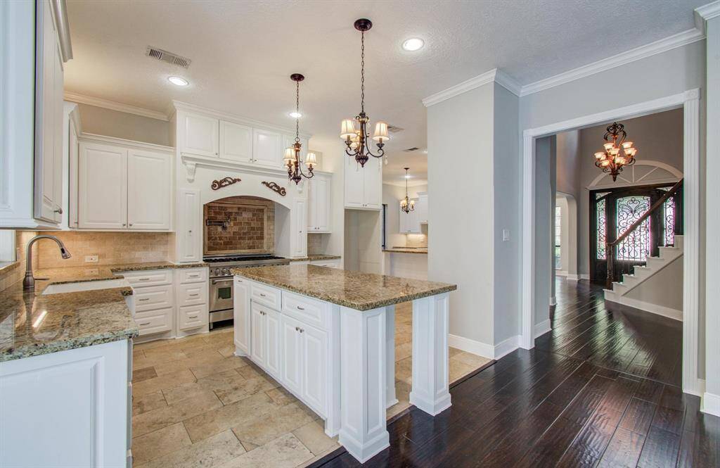 This photo showcases a spacious, elegant kitchen with white cabinetry, granite countertops, and a central island. The room features stylish chandeliers and opens to a hallway with dark hardwood flooring, leading to a staircase with decorative ironwork.
