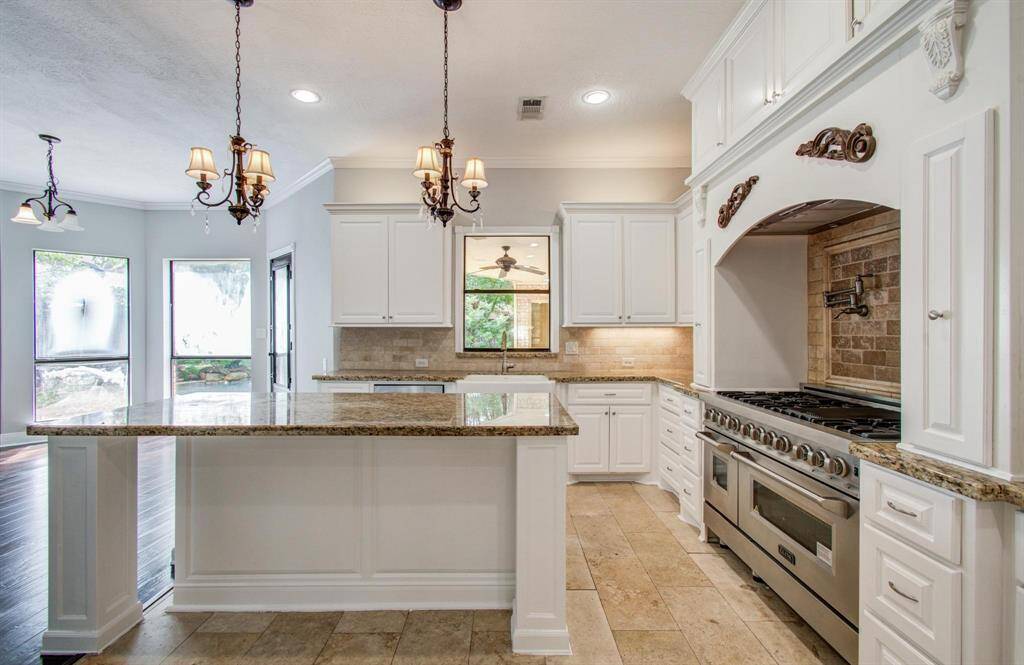 This spacious kitchen features elegant white cabinetry, granite countertops, and a large island. It has a high-end gas stove with a decorative range hood and warm stone backsplash. Natural light pours in through large windows, complementing the sophisticated pendant lighting.