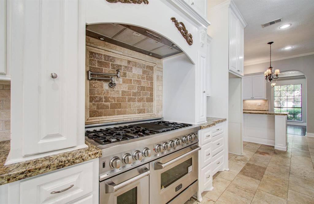 This kitchen features a large stainless steel gas range with a decorative hood and a tile backsplash. It has granite countertops, white cabinetry, and leads into a bright, open dining area with a chandelier.