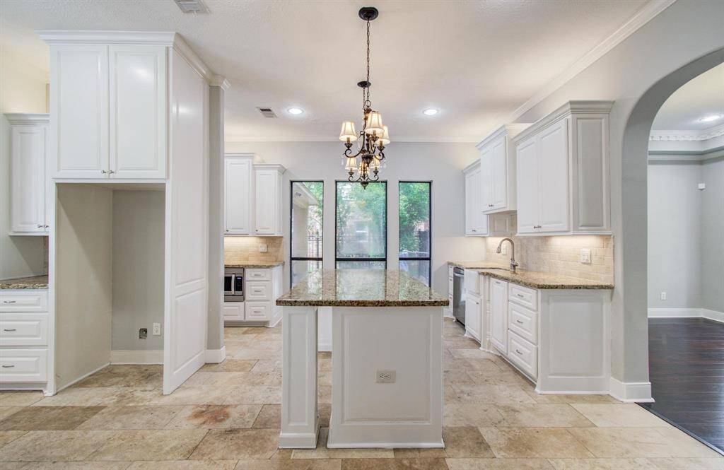 Bright kitchen with white cabinetry, granite countertops, and a central island. Features a tile floor, stainless steel appliances, and a chandelier overhead. Large windows provide natural light, and an open archway connects to additional living space.