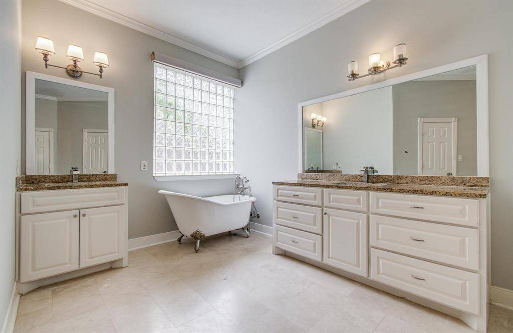 Spacious bathroom featuring dual vanities with granite countertops, a large mirror, elegant light fixtures, and a classic freestanding clawfoot tub. The room is brightened by a glass block window, providing a modern touch and natural light.