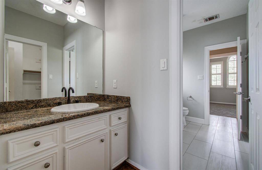 This photo shows a bathroom with a granite countertop and a single sink. It features white cabinetry, a large mirror, and modern lighting. The room connects to a closet and a hallway leading to a carpeted room with large windows.