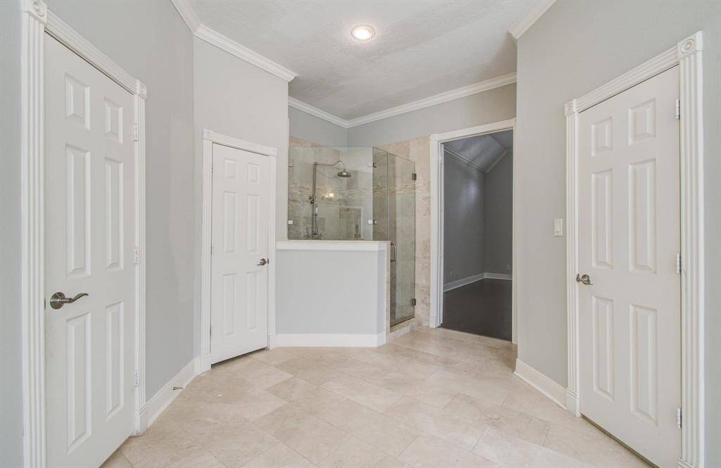 This photo features a spacious bathroom with a glass-enclosed shower, neutral wall colors, and elegant tile flooring. It includes multiple doors, likely leading to closets or additional rooms, and features crown molding for a refined look.