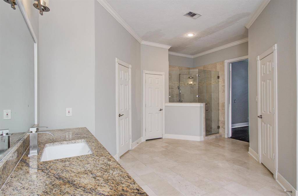 This bathroom features a spacious layout with a granite countertop, large mirror, and ample storage. It includes a glass-enclosed shower, neutral tile flooring, and soft gray walls, creating a modern and clean atmosphere.