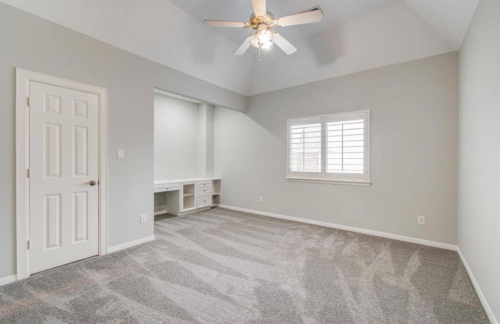 This room features neutral gray walls, a ceiling fan, and plush carpet. It includes a built-in desk with storage, a window with shutters, and a white door. Ideal for a bedroom or home office.