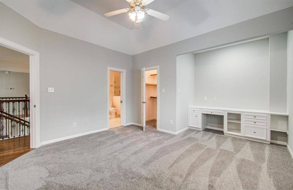 This room features a spacious layout with soft gray carpeting and walls, a ceiling fan, and built-in desk space. It includes access to a bathroom and closet, and overlooks a hallway with elegant railing.