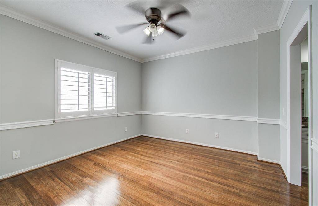 This room features polished hardwood floors, soft gray walls with white trim, and a ceiling fan. The window has plantation shutters, allowing natural light to brighten the space.
