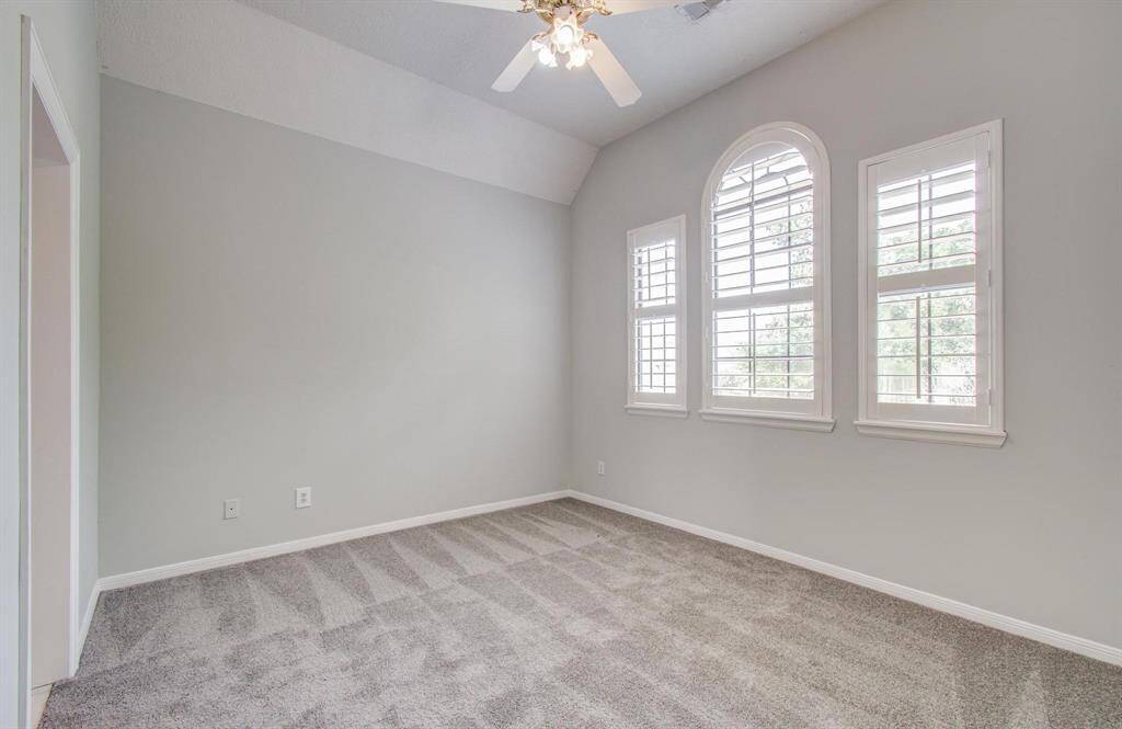Bright bedroom with vaulted ceiling, neutral walls, and plush carpet. Features large arched windows with shutters and a ceiling fan.