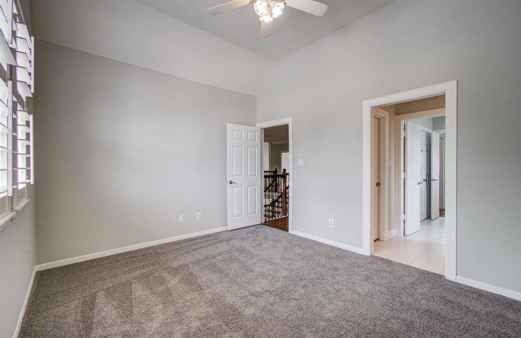 This room features soft gray walls and carpet, a ceiling fan, and ample natural light through windows with shutters. It connects to a hallway and a tiled area, offering versatile space for various uses.