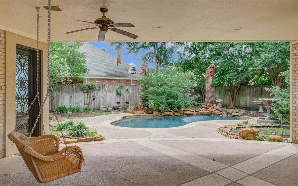 This backyard oasis features a covered patio with a ceiling fan and a charming wicker swing. It overlooks a serene pool surrounded by lush landscaping and a stone fountain, perfect for relaxation and outdoor entertainment.