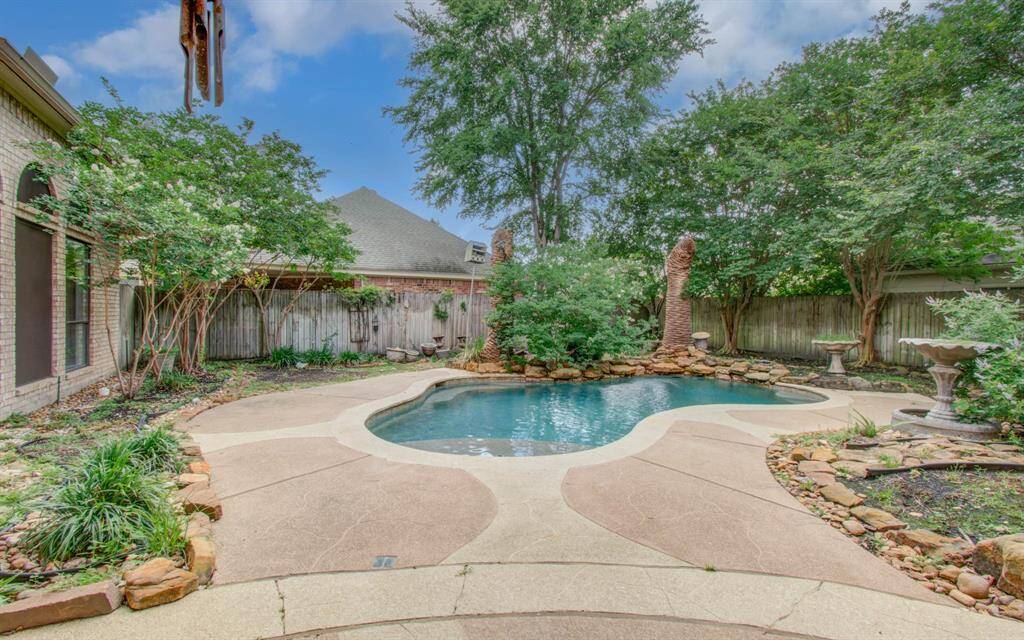 Charming backyard oasis featuring a curvy pool surrounded by lush greenery and mature trees. The stone accents and spacious patio area create a serene environment, perfect for relaxation and entertaining.