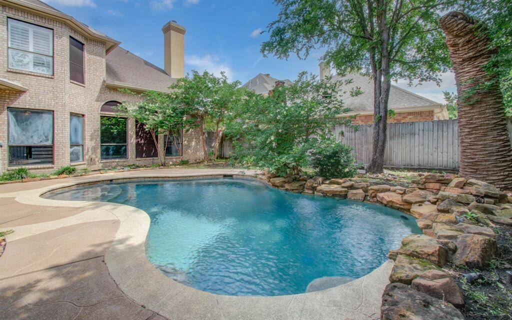 This photo showcases a spacious backyard with a beautifully landscaped pool, surrounded by stone accents and lush greenery, adjacent to a brick home. Perfect for relaxation and entertaining.