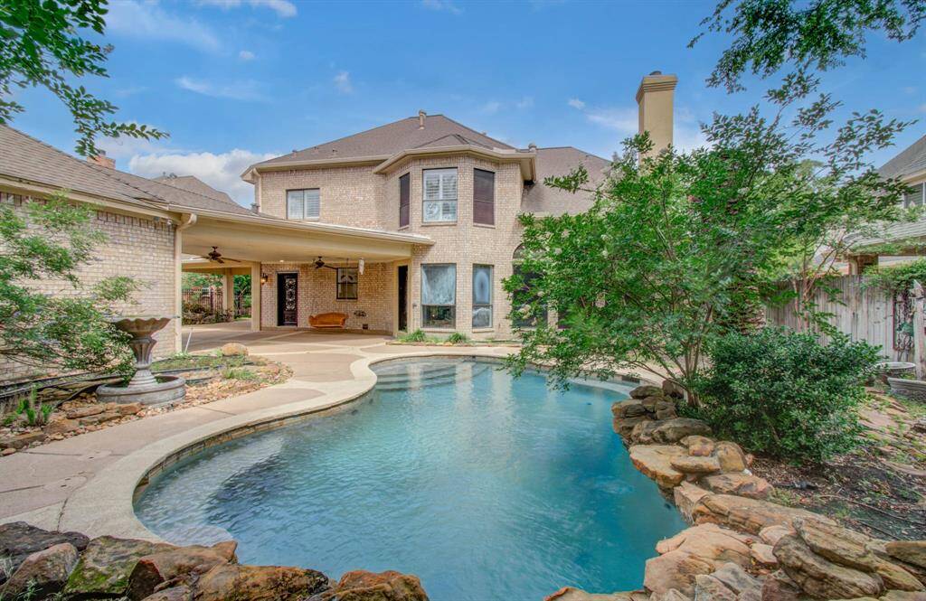 This inviting backyard features a beautiful curved pool surrounded by lush landscaping and a stone border. A covered patio with ceiling fans provides a perfect spot for relaxation and entertainment. The brick exterior of the home adds a classic touch to this serene outdoor space.