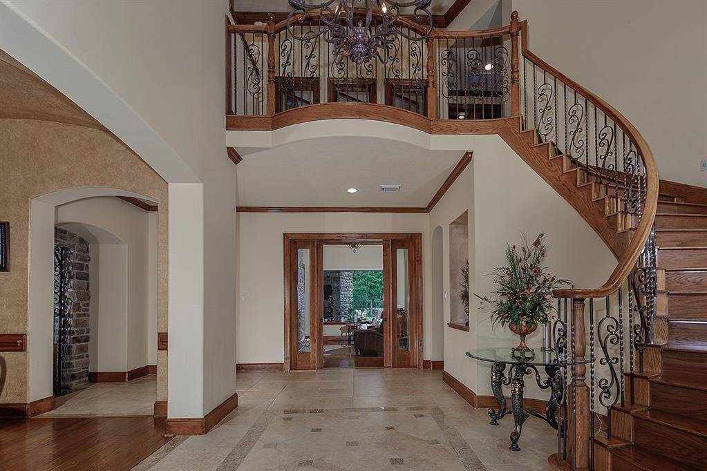 Impressive foyer with elegant flooring and a beautiful curved wooden staircase with intricate ironwork. The space is enhanced by a high ceiling and a striking chandelier creating a welcoming and spacious atmosphere.