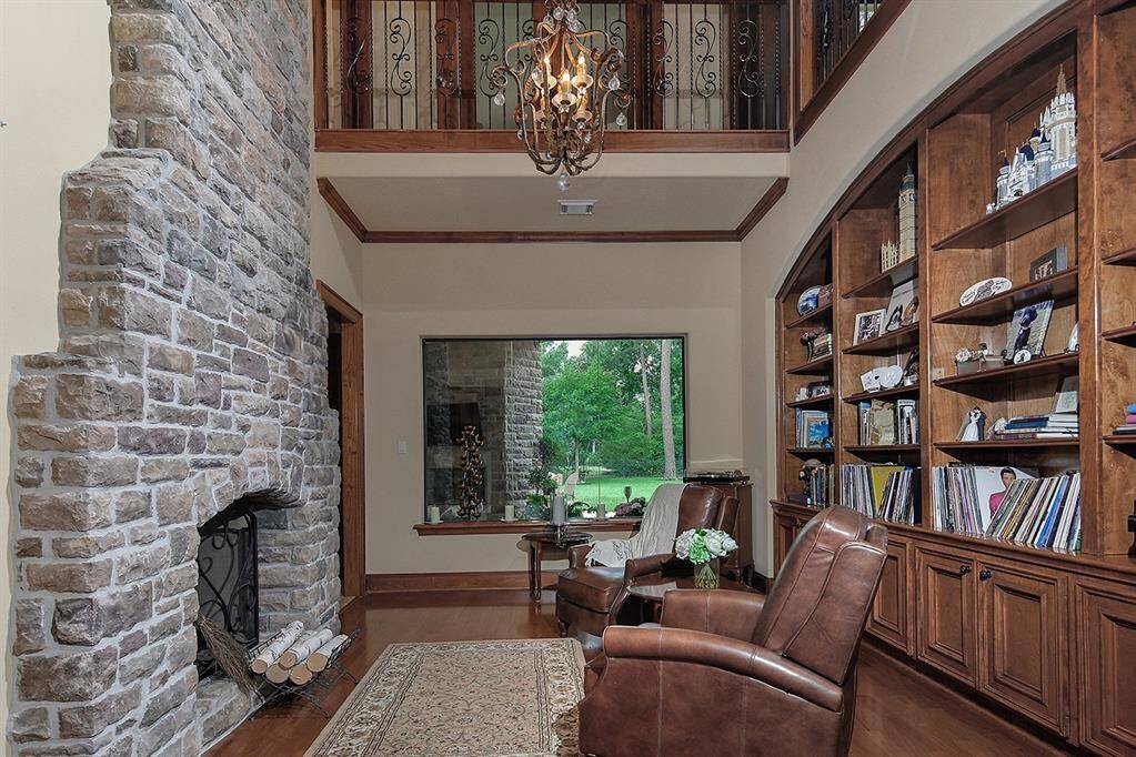 Impressive living room/library/den features a charming stone fireplace, custom built-in bookshelves, and a large picturesque window. A chandelier and upper-level railing add sophistication to this space.
