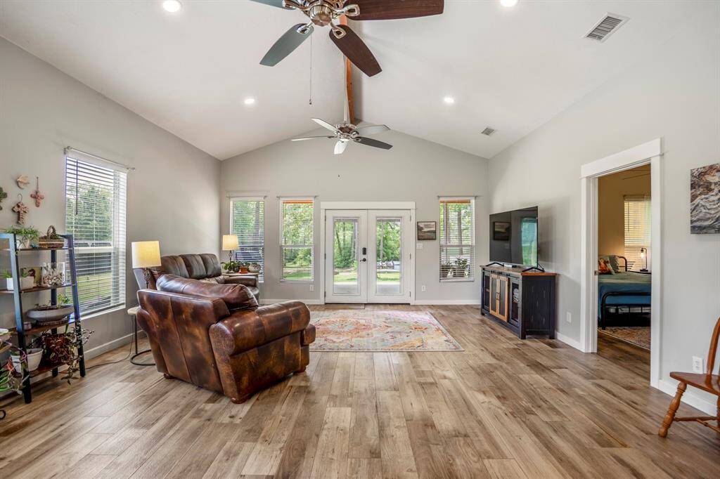 Bright and spacious living room with vaulted ceilings, and large windows. Features double doors opening to a lush outdoor view, cozy seating, and modern ceiling fans.