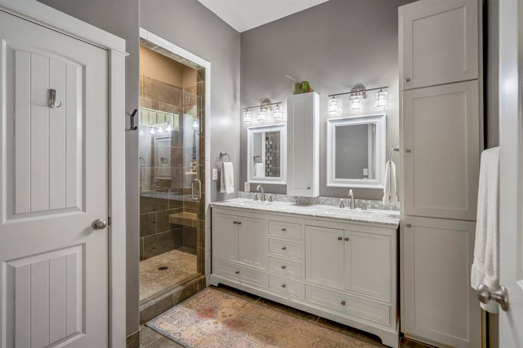 This primary bathroom is complete with a double vanity and marble countertop, ample storage, and stylish lighting fixtures. It includes a spacious, tiled walk-in shower with glass doors.