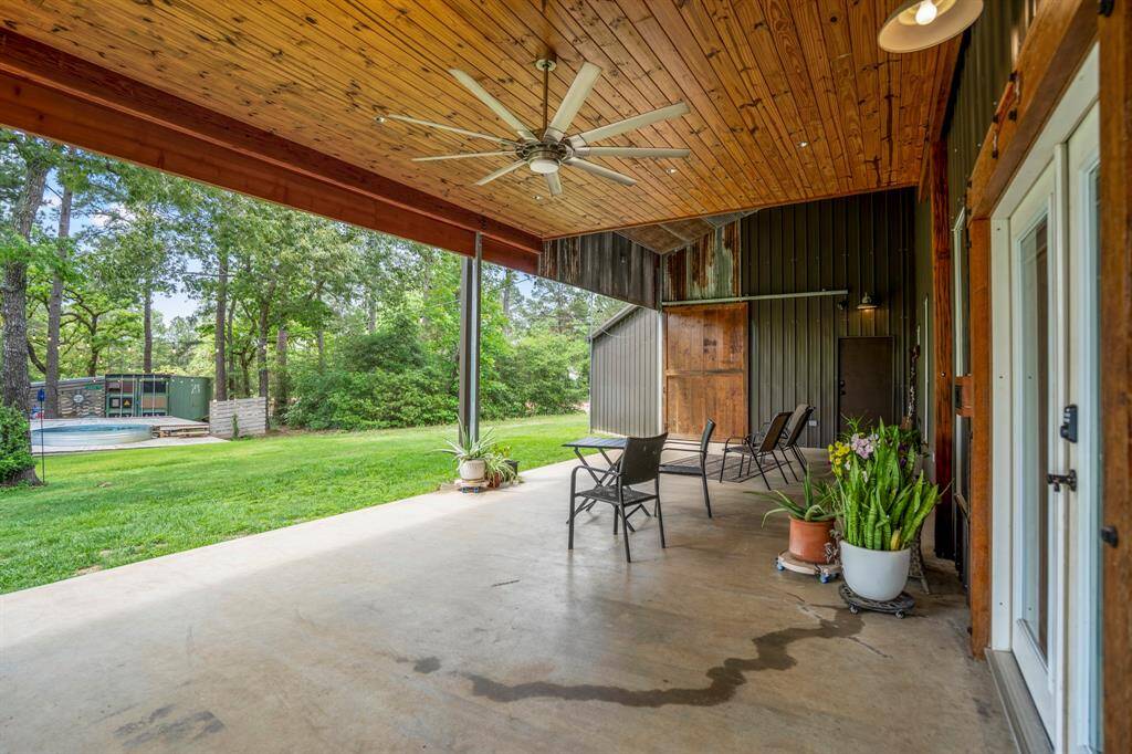 Spacious covered patio with recessed lighting and a ceiling fan, overlooking a lush backyard with tall trees. The are is perfect for outdoor entertaining, leading to a pool surrounded by a wooden deck.