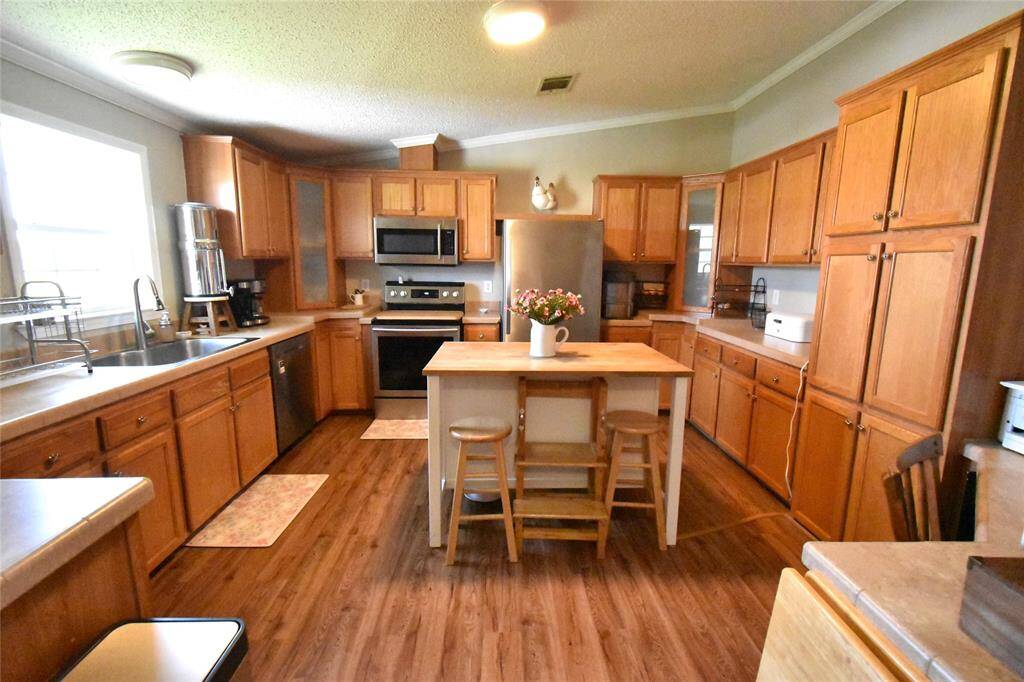 This spacious kitchen features ample wooden cabinetry, stainless steel appliances, and a central island with seating. Large windows provide plenty of natural light, enhancing the warm, inviting atmosphere. Perfect for cooking and entertaining.