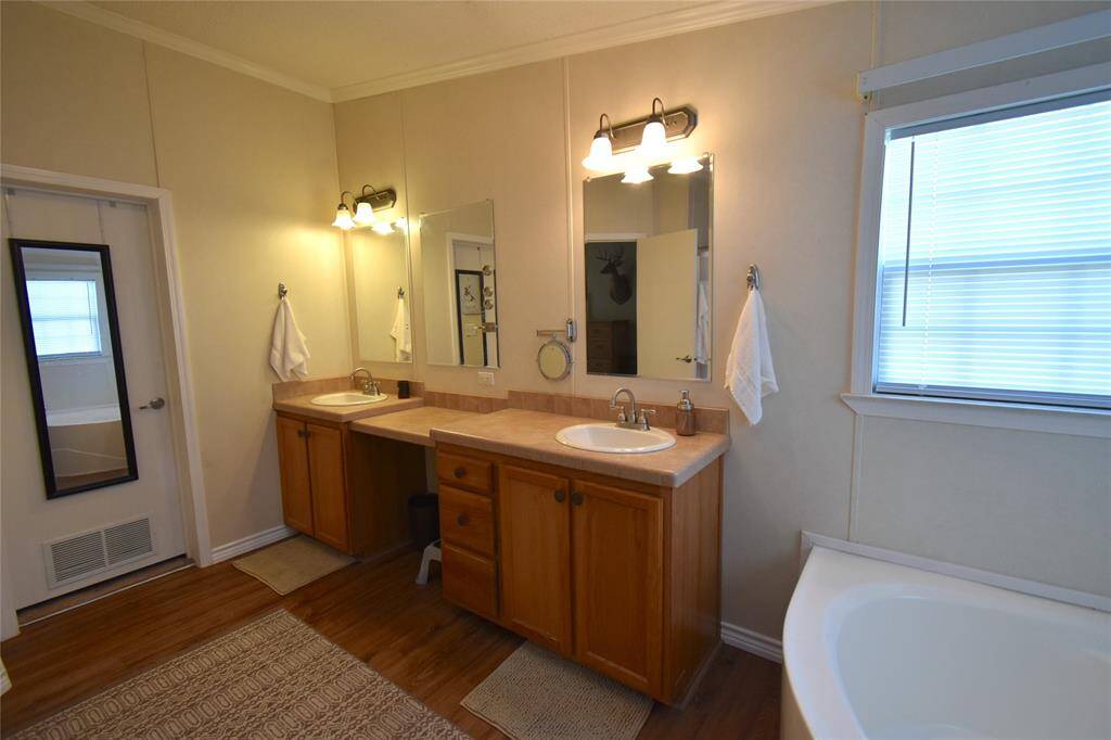 This bathroom features a double vanity with wooden cabinets, large mirrors, and elegant lighting. It includes a soaking tub, a window with blinds for natural light, and wood-style flooring, creating a warm and inviting space.