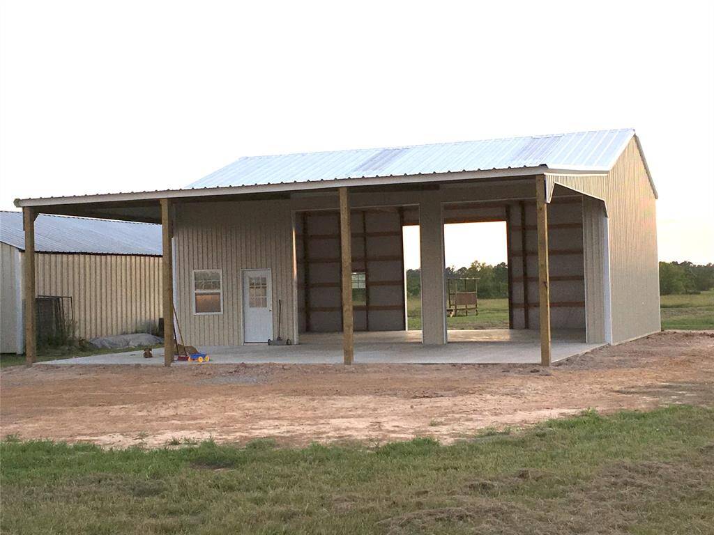 This photo shows a metal barn or workshop with an open front, supported by wooden posts. It has a white door and window on the left side, with a clear, spacious interior. The building is set on a concrete slab, surrounded by a grassy area, ideal for storage or projects in a rural setting.