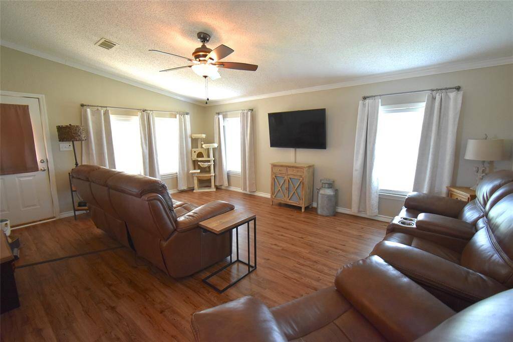 This spacious living room features beautiful flooring, a ceiling fan, and ample natural light from large windows. It includes comfortable seating, a wall-mounted TV, and a cozy, neutral color palette, perfect for relaxation and entertainment.