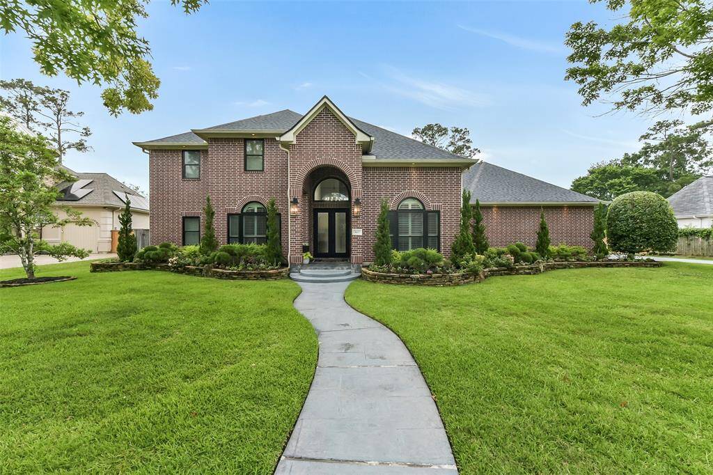 This photo shows a charming two-story brick home with a well-maintained front yard. It features a welcoming pathway leading to the entrance, surrounded by lush greenery and landscaped garden beds. The house has a classic design with large windows and a gabled roof.