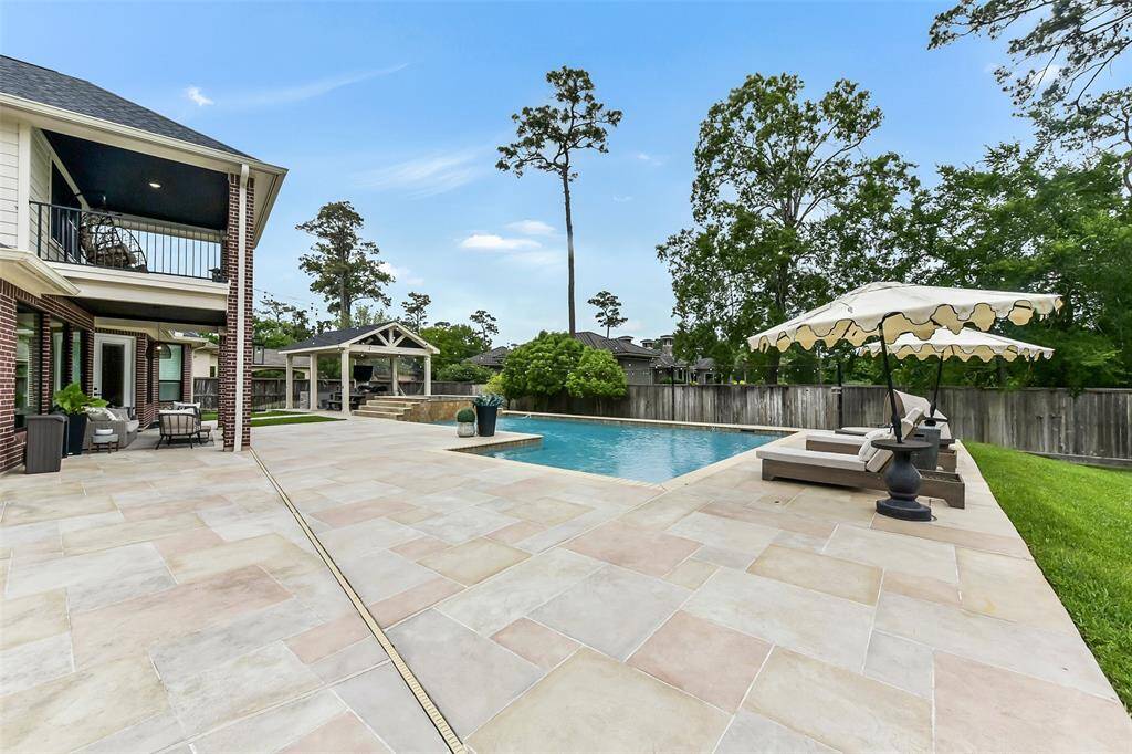 This property features a spacious backyard with a large swimming pool, surrounded by a tiled like carvestone patio floors.There's a covered seating area, perfect for outdoor relaxation. The yard is bordered by tall trees, providing privacy and a natural backdrop.