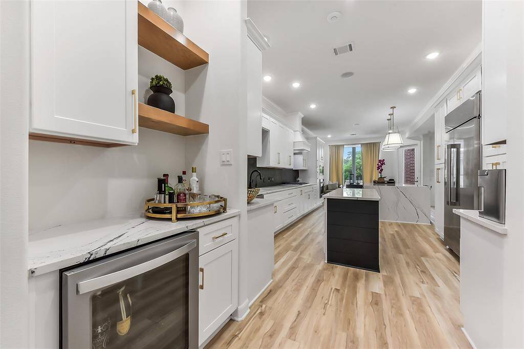 This modern kitchen features sleek white cabinetry, a large island with a waterfall countertop, and stainless steel appliances. The open layout is complemented by wood flooring and recessed lighting, leading to a bright dining area with large windows.