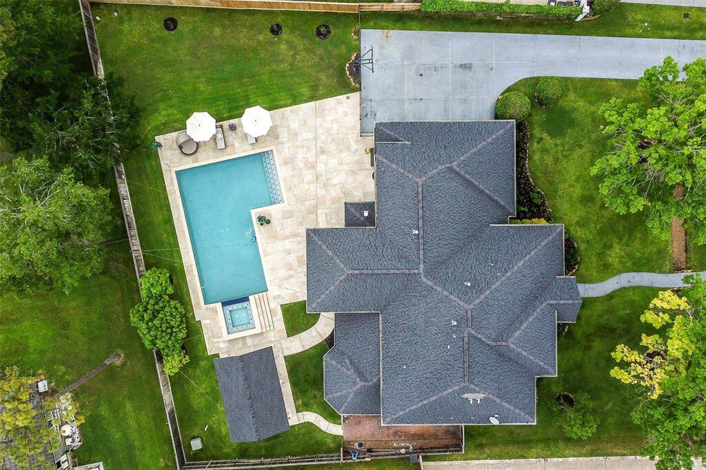 This aerial view showcases a spacious property with a large, modern home featuring a dark roof and an inviting backyard. The outdoor area includes a sleek pool with a hot tub, surrounded by a stone patio and greenery, perfect for relaxation and entertaining. The driveway provides ample parking space, and the property is enclosed by lush trees, offering privacy and tranquility.