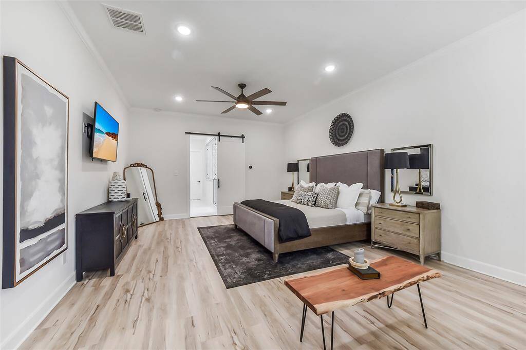 Modern bedroom with neutral tones, featuring a stylish ceiling fan, wood flooring, and a sliding barn door leads to an ensuite bathroom.