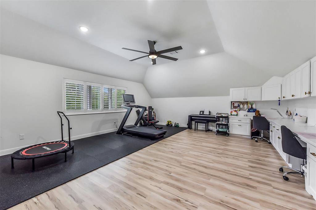 This spacious, versatile room features a modern design with light wood flooring and recessed lighting. It includes a home gym area with a treadmill and mini trampoline, a work or study space with built-in desks and cabinets, and a ceiling fan for comfort. Ideal for fitness enthusiasts or those needing a functional workspace.