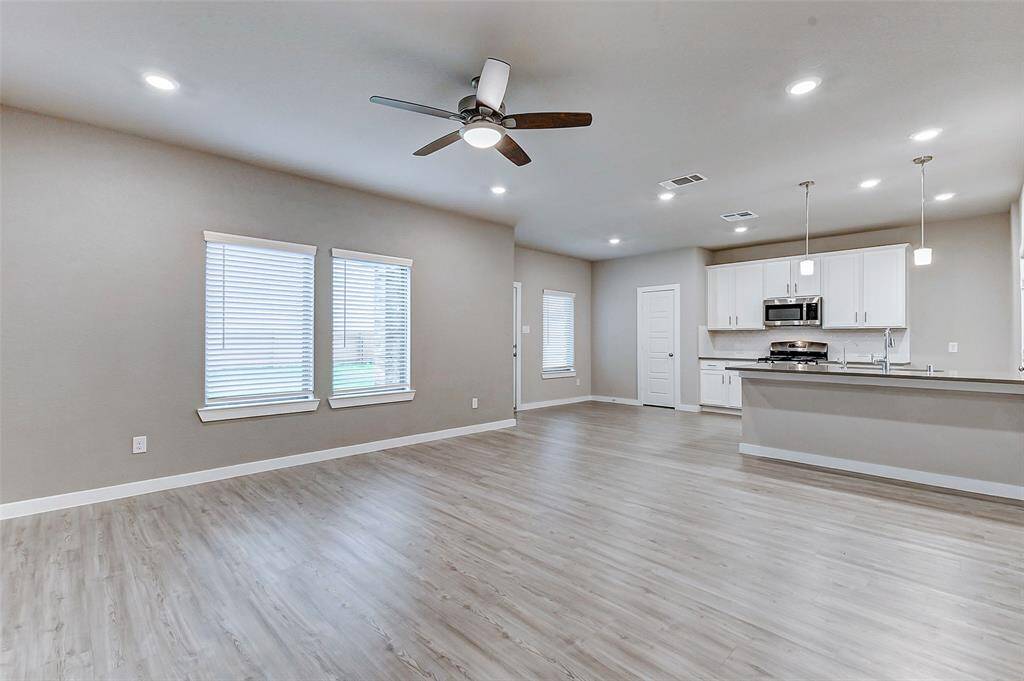 The spacious living room exudes a serene ambiance with large windows inviting natural light. Stylish wood-like laminate floors pair perfectly with lush greenery accents and modern recessed lighting. A ceiling fan adds comfort, while the space is ideal for entertaining or unwinding in peaceful elegance.