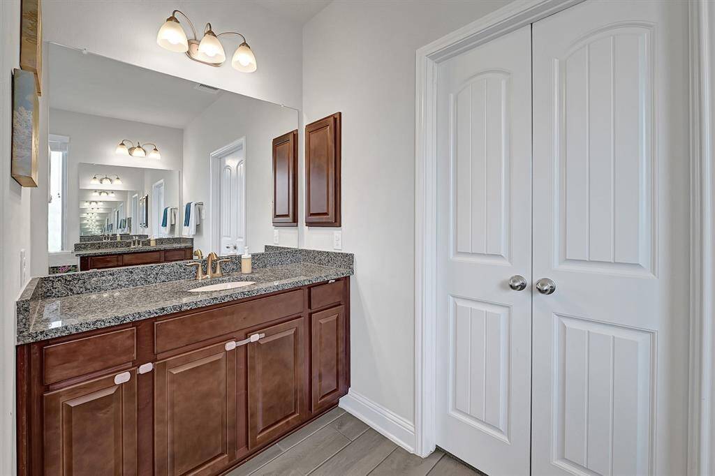 Dual sinks, grey wood-look tile flooring, Nutmeg cabinetry, and gold fixtures line your private ensuite bathroom.