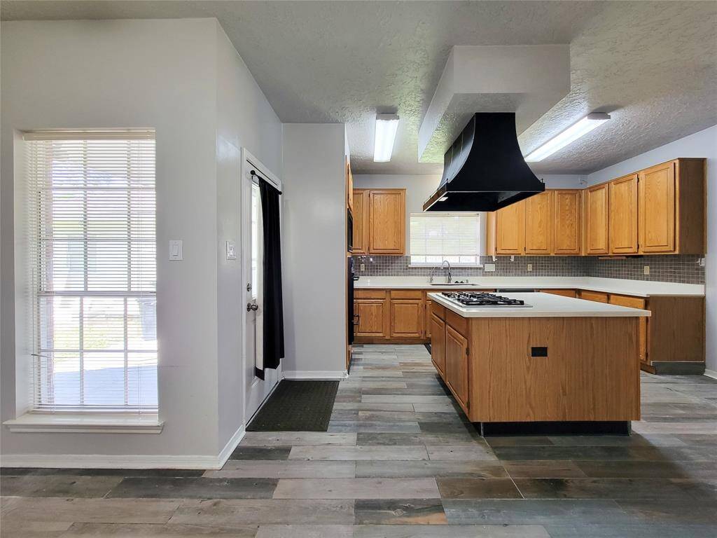Spacious kitchen with modern lighting, abundant natural light, and sleek tile flooring. Features ample cabinetry and a large island with a cooktop, perfect for culinary enthusiasts.