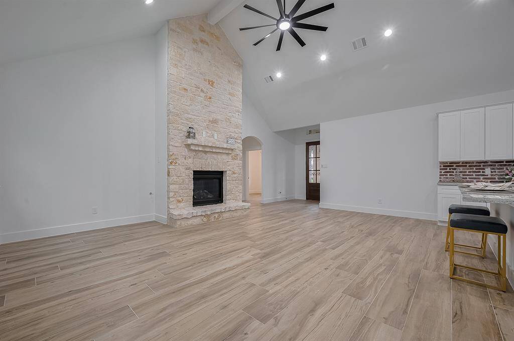This spacious living area features high vaulted ceilings, a modern ceiling fan, and a cozy stone fireplace. The open layout includes a sleek kitchen with a brick backsplash and a breakfast bar with seating, perfect for entertaining. Light wood flooring and neutral walls create a bright, inviting atmosphere.