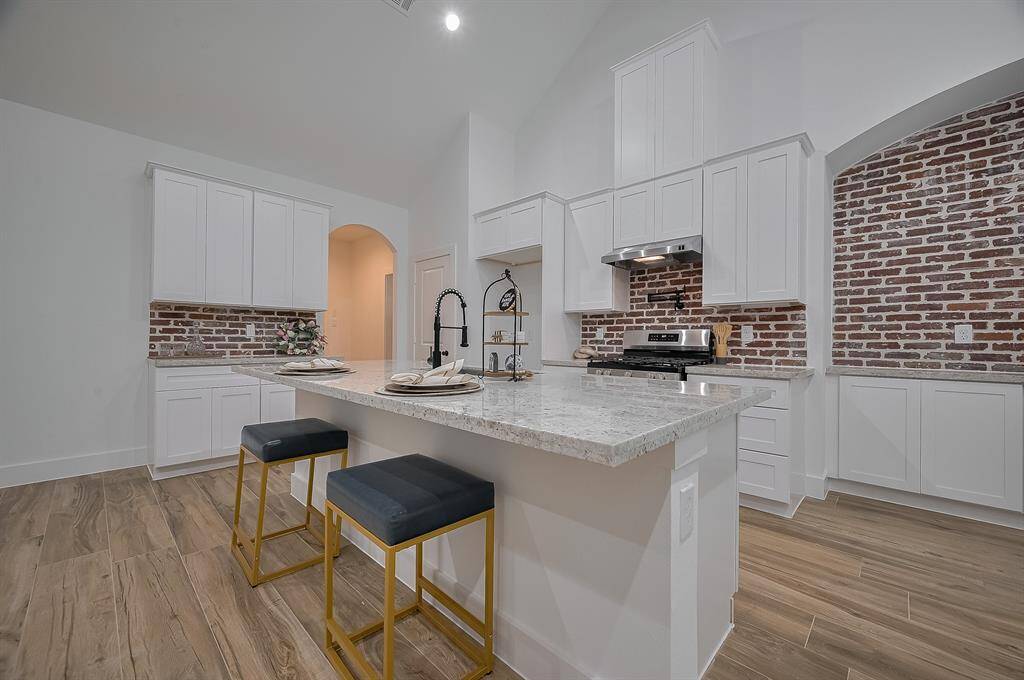 This modern kitchen features sleek white cabinetry and a stylish brick backsplash. It includes a spacious island with a marble countertop, complemented by two elegant bar stools. The stainless steel appliances and wood-look flooring add a touch of sophistication.