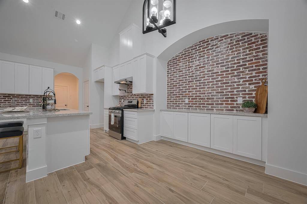 This kitchen features a modern design with white cabinetry, stainless steel appliances, and a large marble island. The exposed brick backsplash adds character, while the light wood flooring and high ceiling create an open, inviting atmosphere.