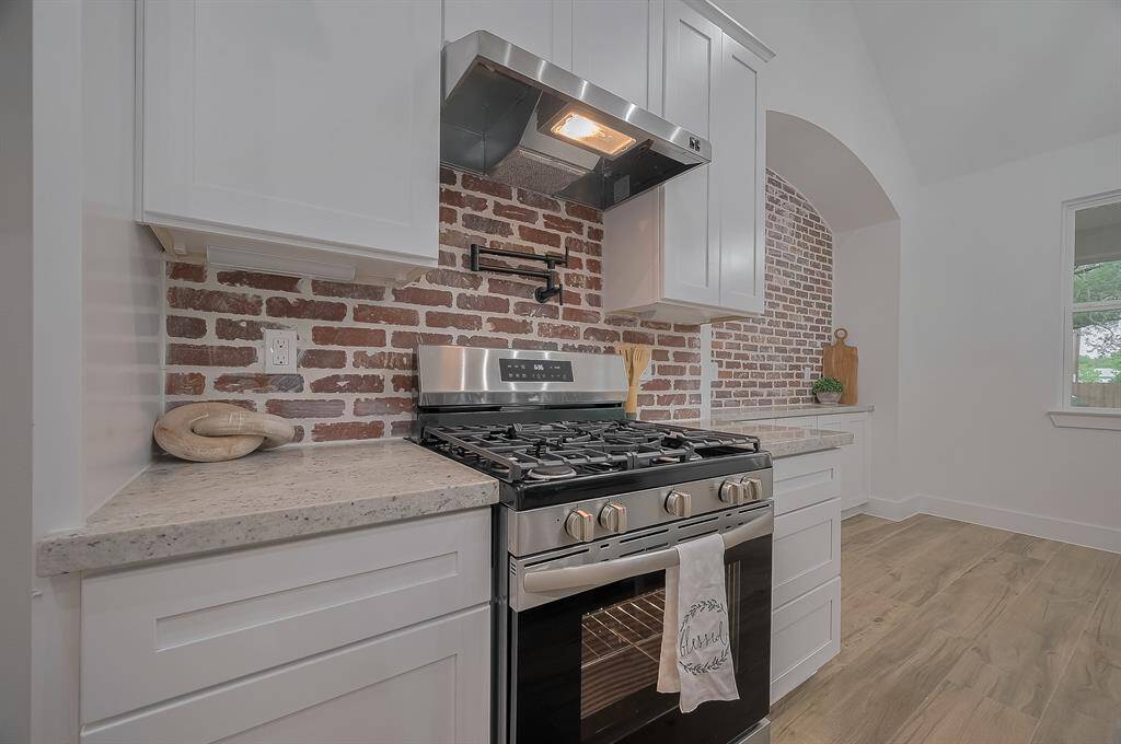 This kitchen features a modern design with a stainless steel gas stove and oven, white cabinetry, and a charming brick backsplash. The space is bright and airy with wood-look flooring and a cozy breakfast nook.