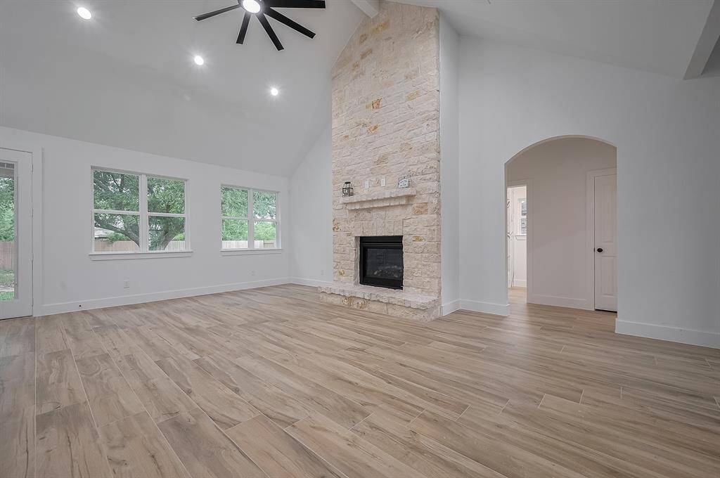 This spacious living room features high vaulted ceilings, a modern ceiling fan, and natural wood-style flooring. The room is bright with large windows and a stone fireplace as a focal point. An arched doorway adds elegance and leads to another area of the home.