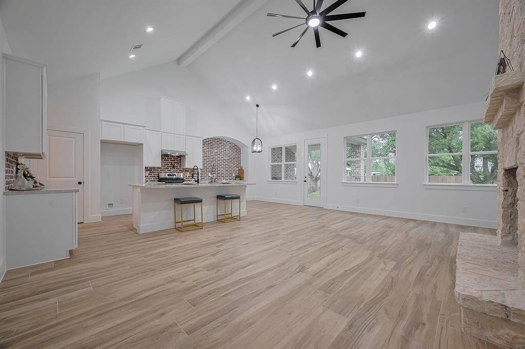 This spacious open-concept living area features high vaulted ceilings, modern recessed lighting, and a ceiling fan. The kitchen boasts an island with seating, stainless steel appliances, and a brick backsplash. Large windows offer plenty of natural light and a view of the backyard, complemented by wood-style flooring and a cozy stone fireplace.