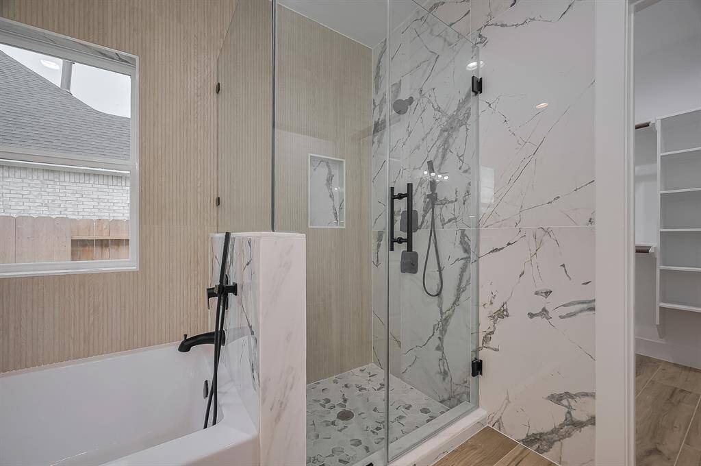 This bathroom features a modern design with a glass-enclosed shower showcasing elegant marble-style tiles. It includes a freestanding bathtub with a sleek black faucet, wood-textured wall accents, and ample natural light from a large window. Perfect for a luxurious and relaxing experience.