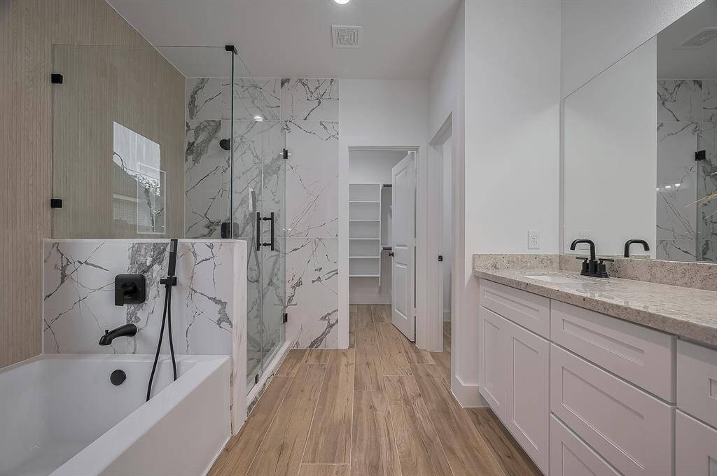 This modern bathroom features a spacious design with a sleek glass-enclosed shower and a luxurious marble-patterned tub. It includes a double vanity with granite countertops, ample storage, and elegant fixtures. The wood-style flooring adds warmth and sophistication.